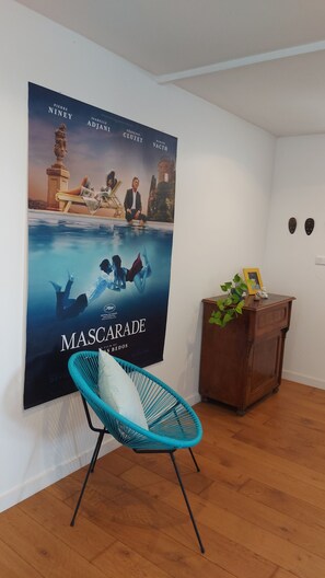 Ein Zimmer mit einem 'MascaraDE'-Poster an der Wand, einem blauen Stuhl, einem Holzschrank und einer kleinen Pflanze.