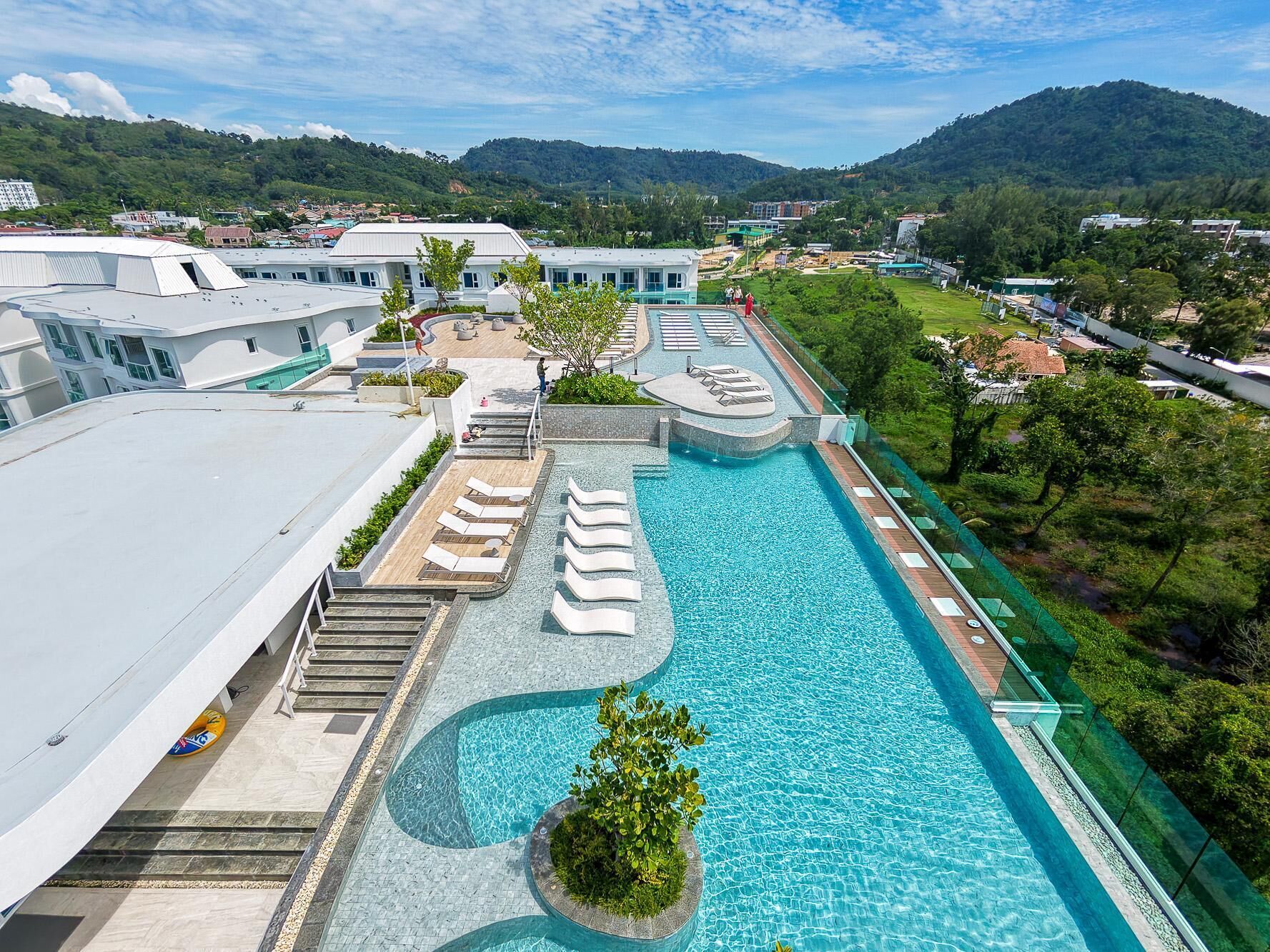 A rooftop swimming pool with lounge chairs, a view of the surrounding landscape, and a building with a white facade.