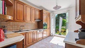 A kitchen with wooden cabinets, a tiled backsplash, a built-in oven, and a large window with a view of greenery outside. - Villa Harmonie - Welkeys (Villeneuve-Loubet)