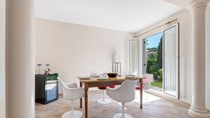 A dining area with a table set for four, white chairs, a black sideboard, and a view through large sliding glass doors. - Villa Harmonie - Welkeys (Villeneuve-Loubet)