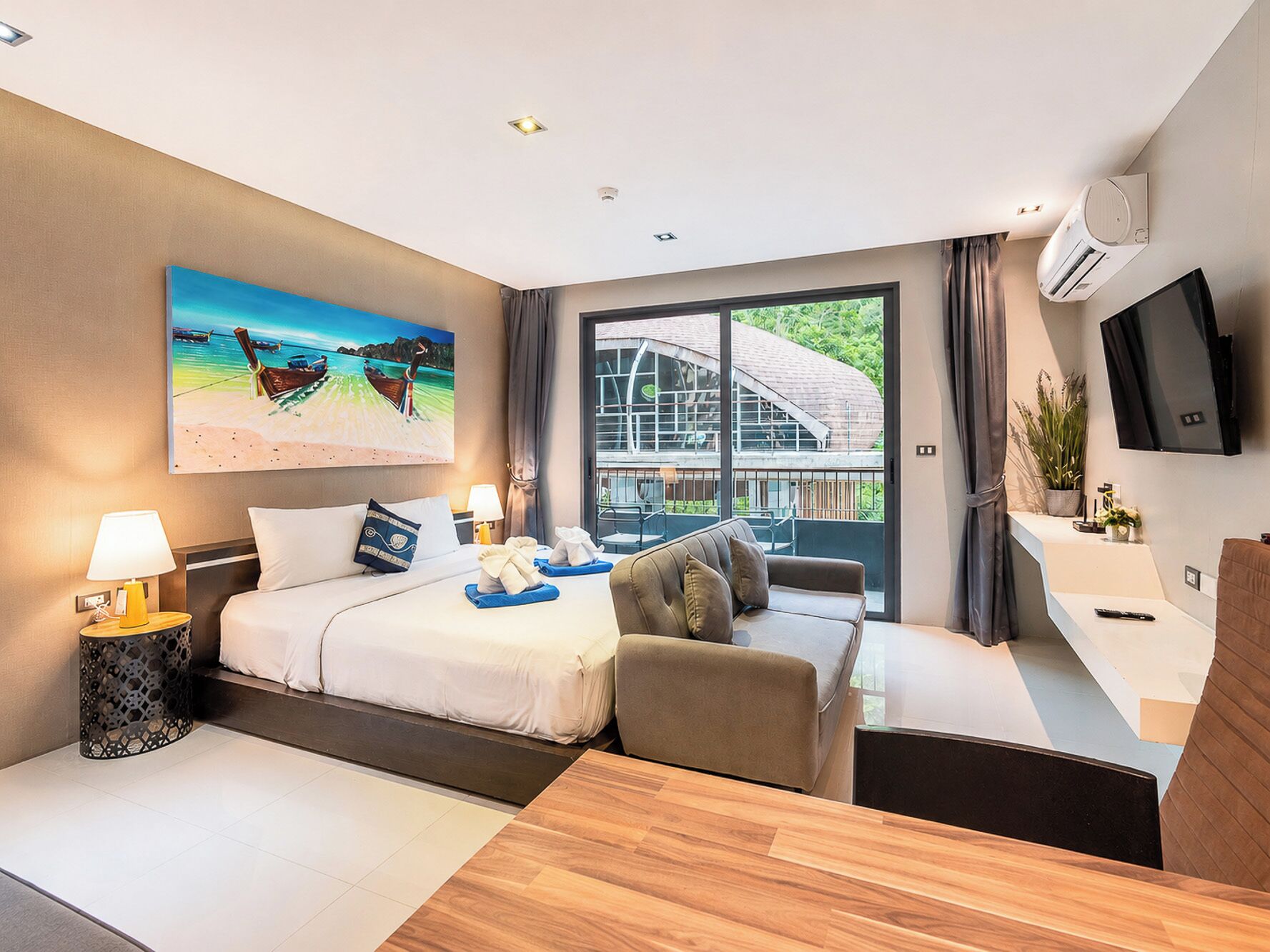 A modern hotel room with a bed, a sofa, a desk, a TV, and a large window with a view of a building and greenery.