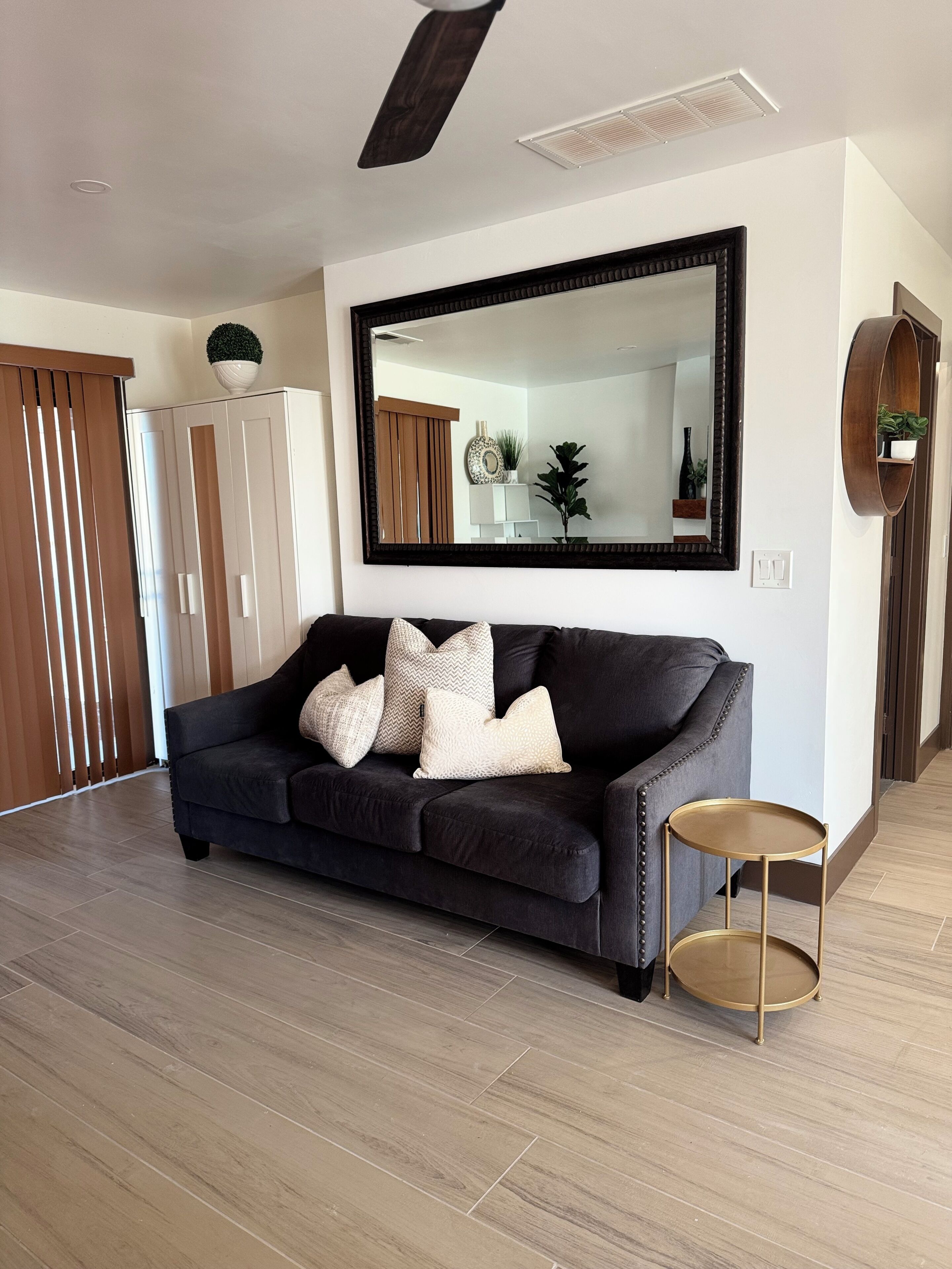 A modern living room with a dark sofa, a round gold side table, a large mirror, and wooden flooring.