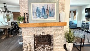 Una sala de estar con chimenea de piedra, un cuadro enmarco de botellas, repisa de madera y alfombra estampada.