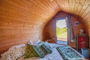 Ein gemütliches Holzhütten-Zimmer mit Bett, Kissen, einem Sessel und einem kleinen Tisch mit Lampe.