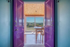 Blick durch eine violette Tür in einen Raum mit Holzbalkendecke, einem Tisch und Stühlen, mit Blick auf eine Landschaft.