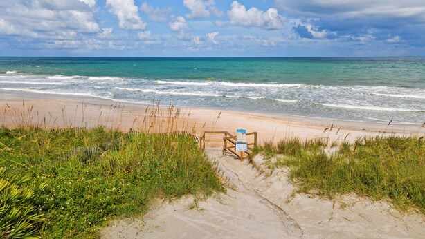 Une plage bordée d’une clôture en bois et agrémentée d’un panneau, entourée de sable et d’herbe.