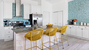 Une cuisine moderne avec des meubles blancs, une cuisinière en acier inoxydable, un four à micro-ondes et un grand îlot central agrémenté de chaises en osier jaune.