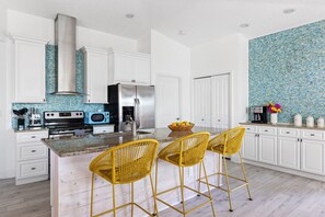 A modern kitchen with white cabinets, a stainless steel range, a microwave, and a large island with yellow wicker chairs. - Flamingo Point | Steps from the Beach (St Augustne)