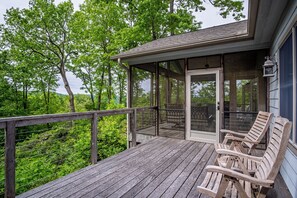 Eine Holzterrasse mit Glasumrandung, Sitzgelegenheiten im Freien und Blick auf einen bewaldeten Bereich.