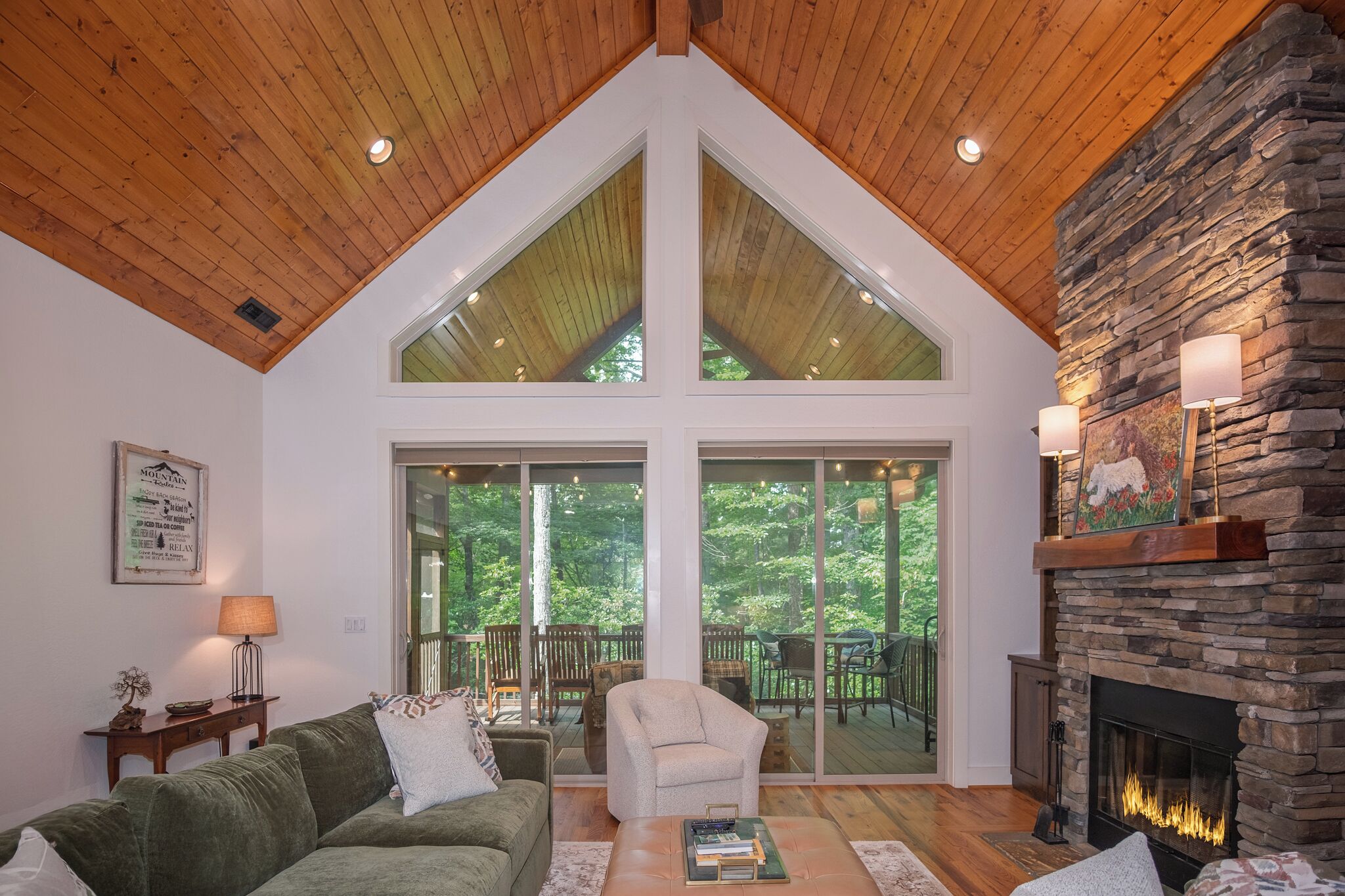 A living room with a stone fireplace, a high wooden ceiling, and large windows overlooking a deck.