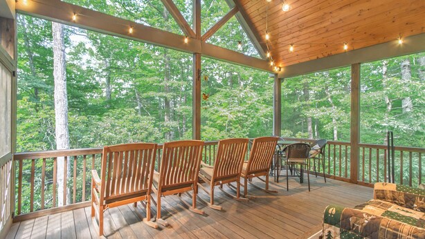 A wooden deck with chairs and a table, surrounded by large windows offering a forest view. - 3BR Retreat in Connestee Falls with Fireplace, Community Pool, and Garage (Brevard)