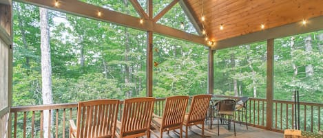 Une terrasse en bois, agrémentée de chaises et d’une table, entourée de grandes fenêtres donnant sur la forêt.