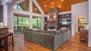 A spacious living room with a large sectional sofa, a stone fireplace, and large windows offering a view of the outdoors. - 3BR Retreat in Connestee Falls with Fireplace, Community Pool, and Garage (Brevard)