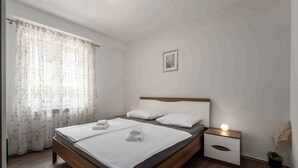 A bedroom with a wooden bed, white bedding, a nightstand with a lamp, and a window with sheer curtains. - Holiday apartment near Pical Beach (Poreč)