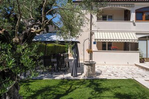 A covered outdoor seating area with tables and chairs, a stone planter with flowers, and a building with a balcony and awning. - Holiday apartment near Pical Beach (Poreč)