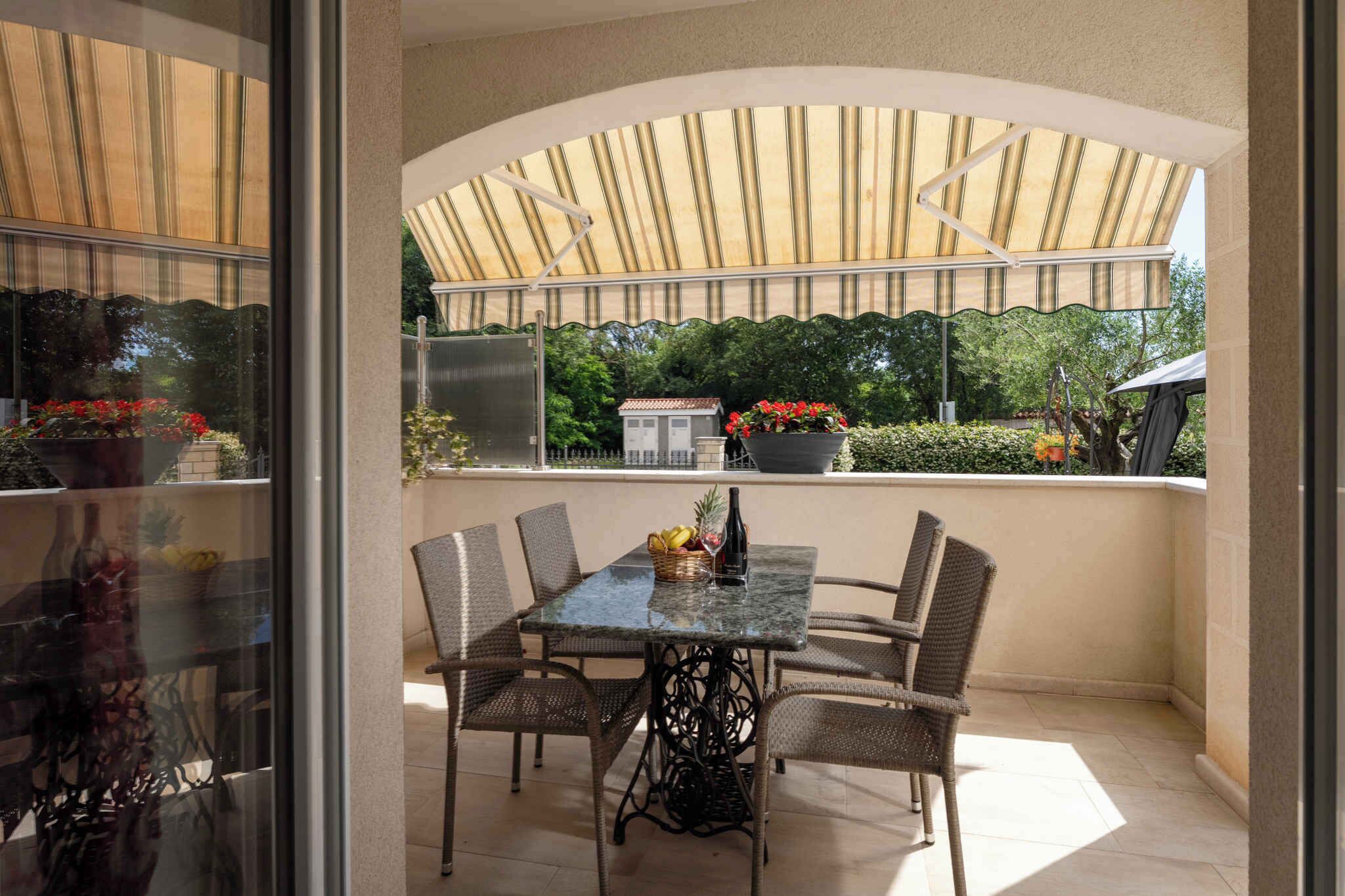 A covered outdoor patio with a table and chairs, a view of greenery, and a building in the background.
