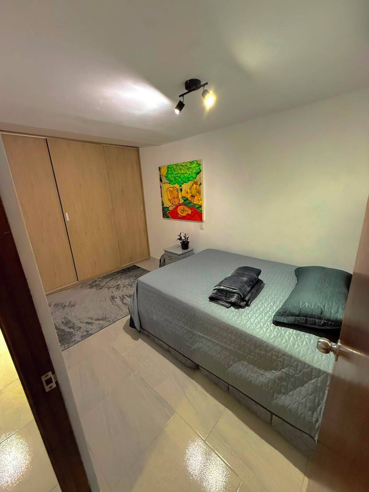 Et soveværelse med seng, garderobe, natbord, et maleri og en loftlampe.