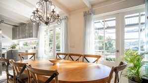A dining area with a round wooden table, six chairs, a chandelier, and large windows with curtains. - Chiara`s Art Cottage 3BR Coastal Garden Retreat (Sorrento)