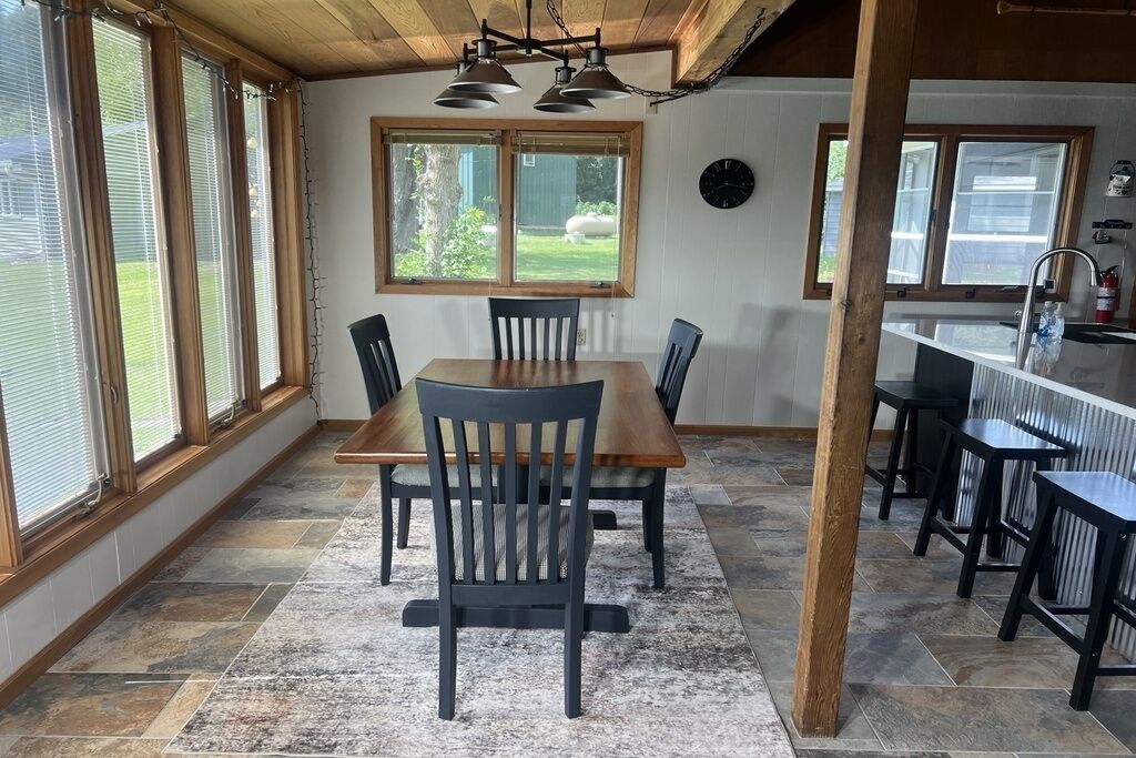 A dining area with a wooden table and chairs, a kitchen with a sink and countertop, and large windows with blinds.