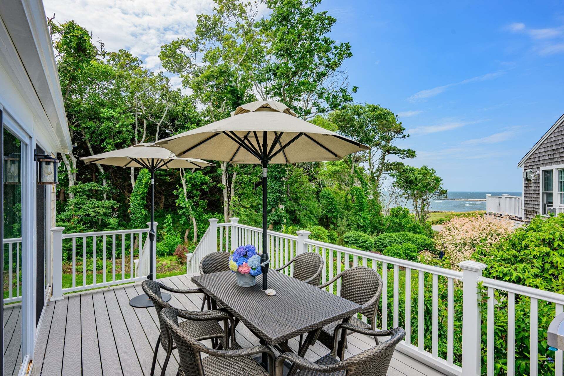 A deck with wicker furniture and a table, surrounded by white railings, overlooking a beach.