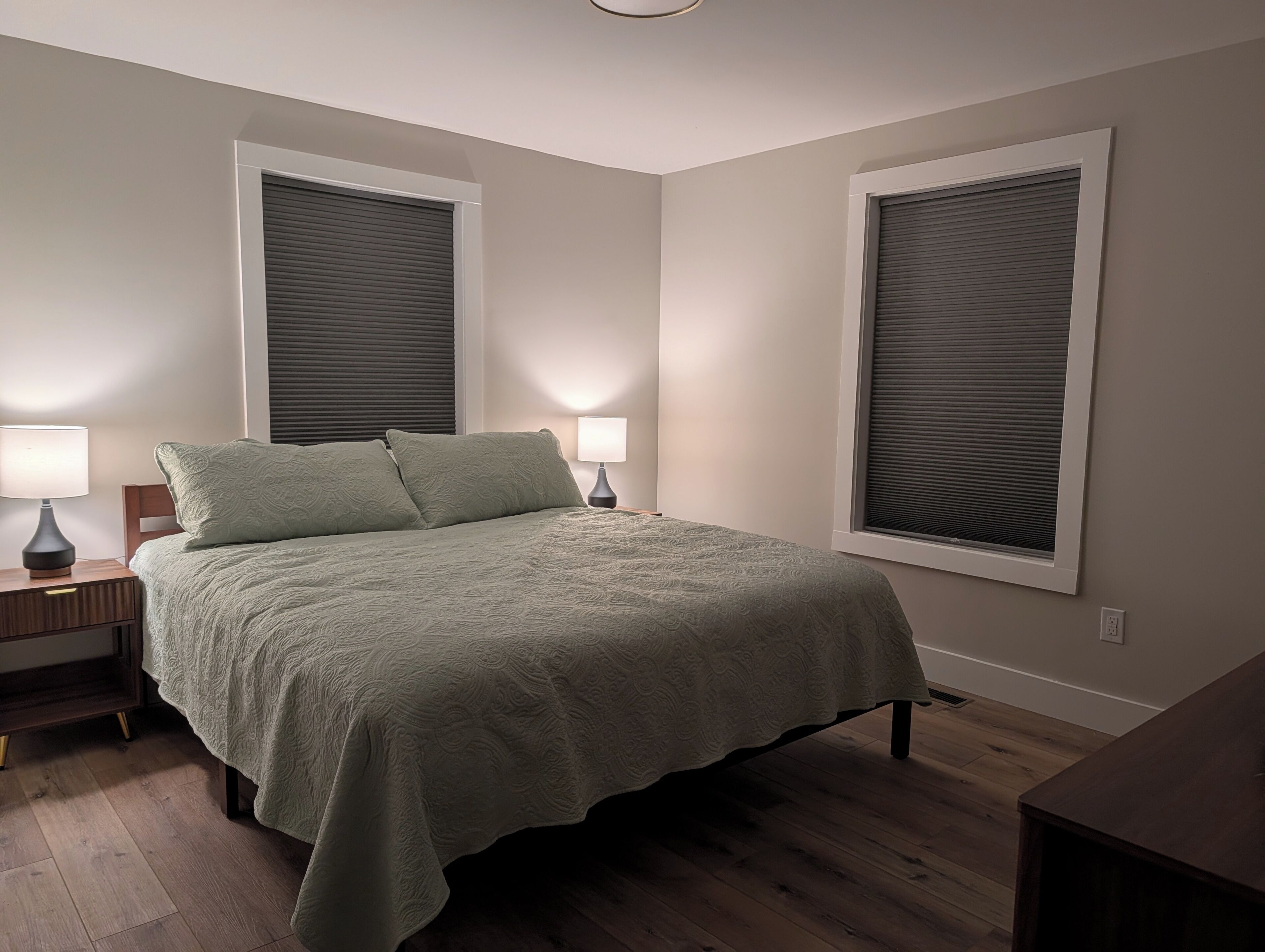 A bedroom with a bed, two bedside lamps, a nightstand, and two windows with blinds.