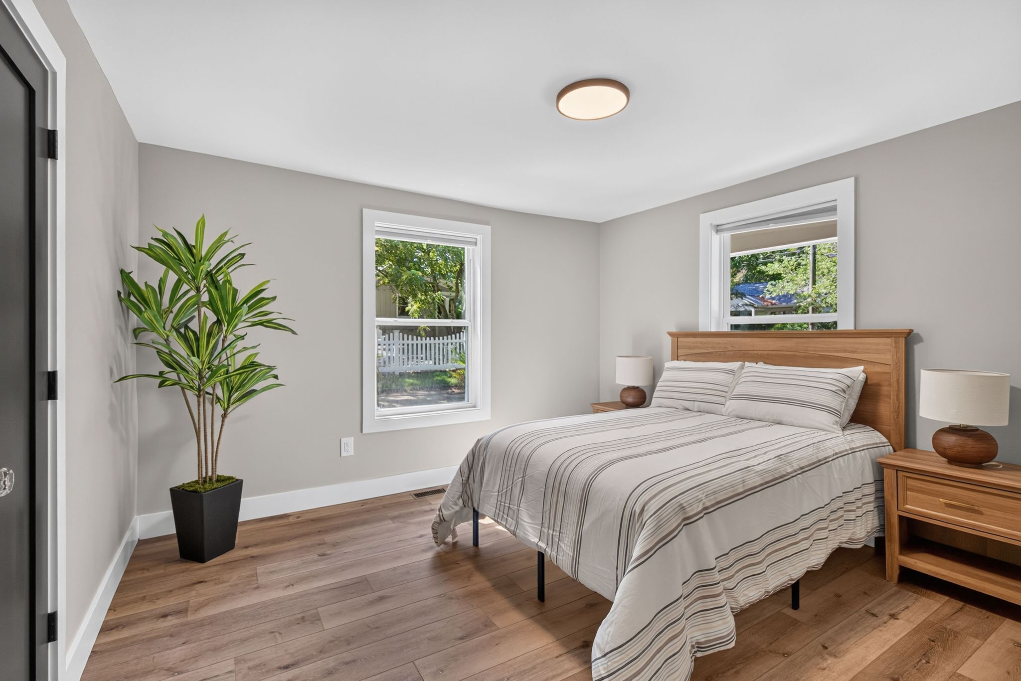 A bedroom with a bed, bedside tables, a potted plant, and windows with views of greenery.