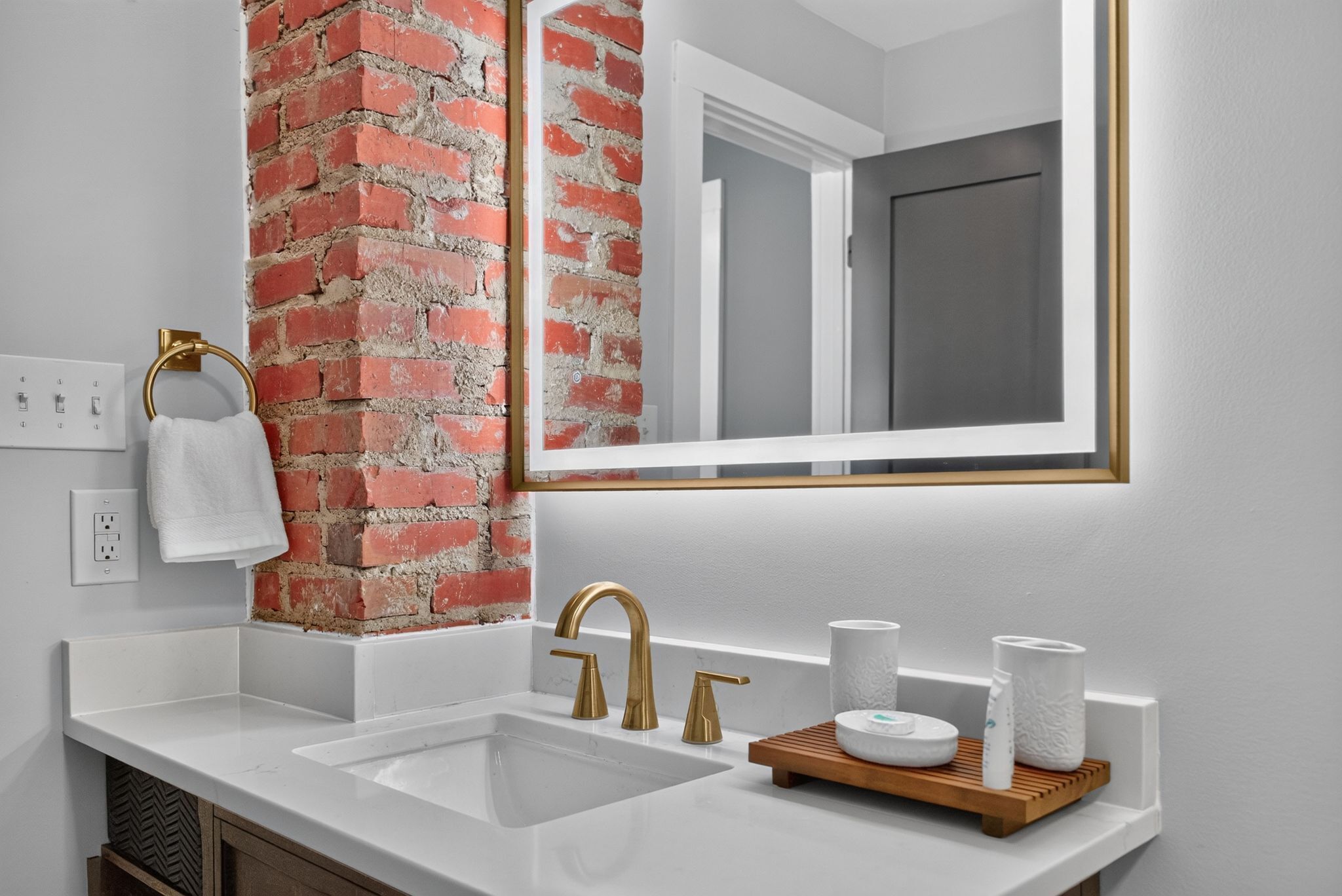A bathroom with a white sink, gold faucets, a mirror, and a brick accent wall.