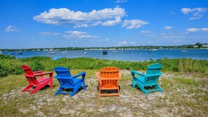 Cuatro sillas Adirondack de colores mirando un cuerpo de agua con barcos y una costa distante.