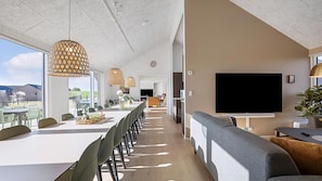 Ein moderner Büroraum mit einem langen Esstisch, mehreren Stühlen, hängenden Pendelleuchten, einem großen Flachbildfernseher und Blick ins Freie.