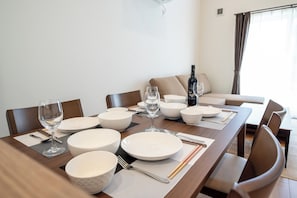 A dining table set with white plates, bowls, cutlery, and a wine glass, with a sofa and curtains in the background. - North Point Kamifurano (Sorachi-gun,Kamifurano-cho)