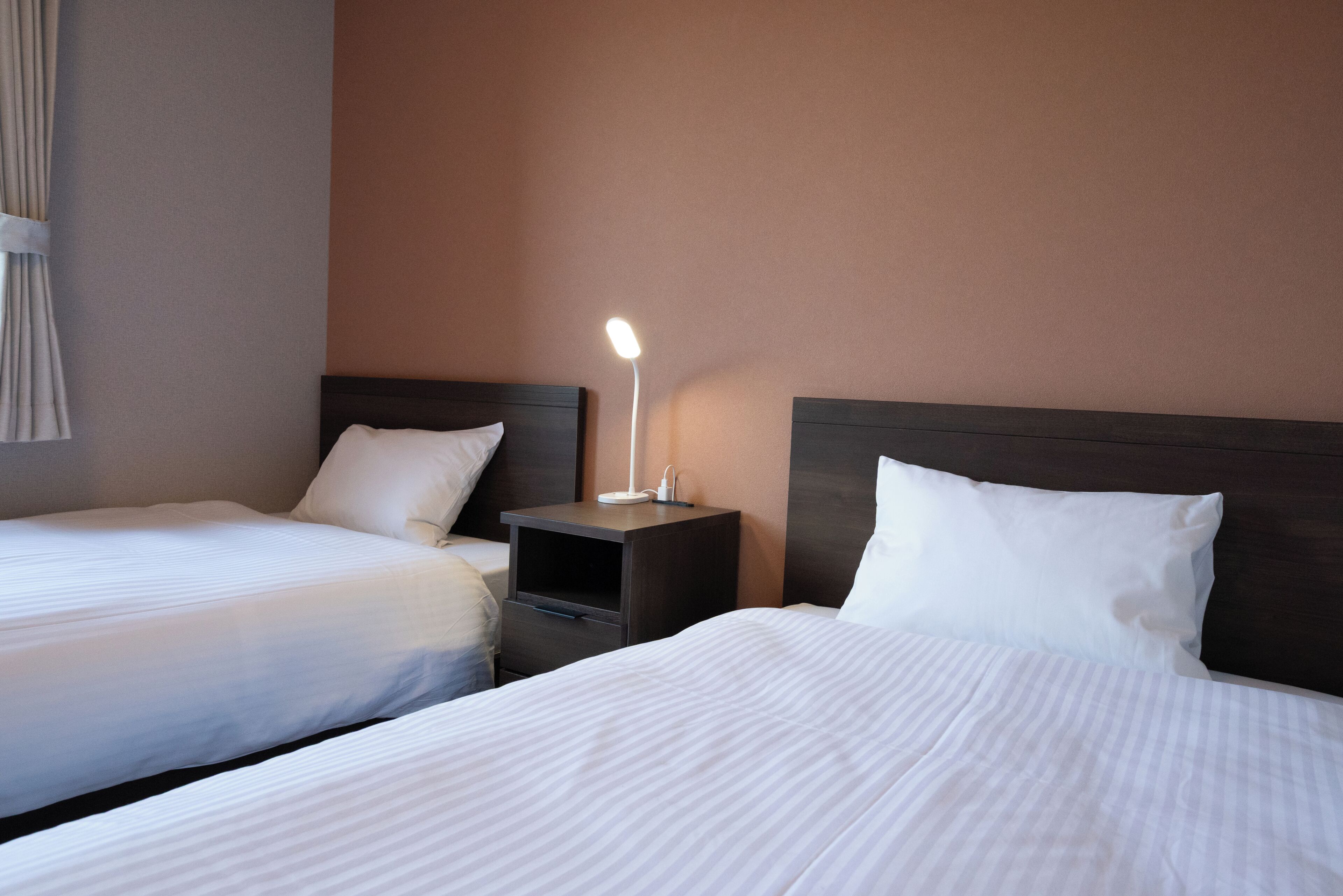 A hotel room with two beds, a nightstand with a lamp, and a window with curtains.
