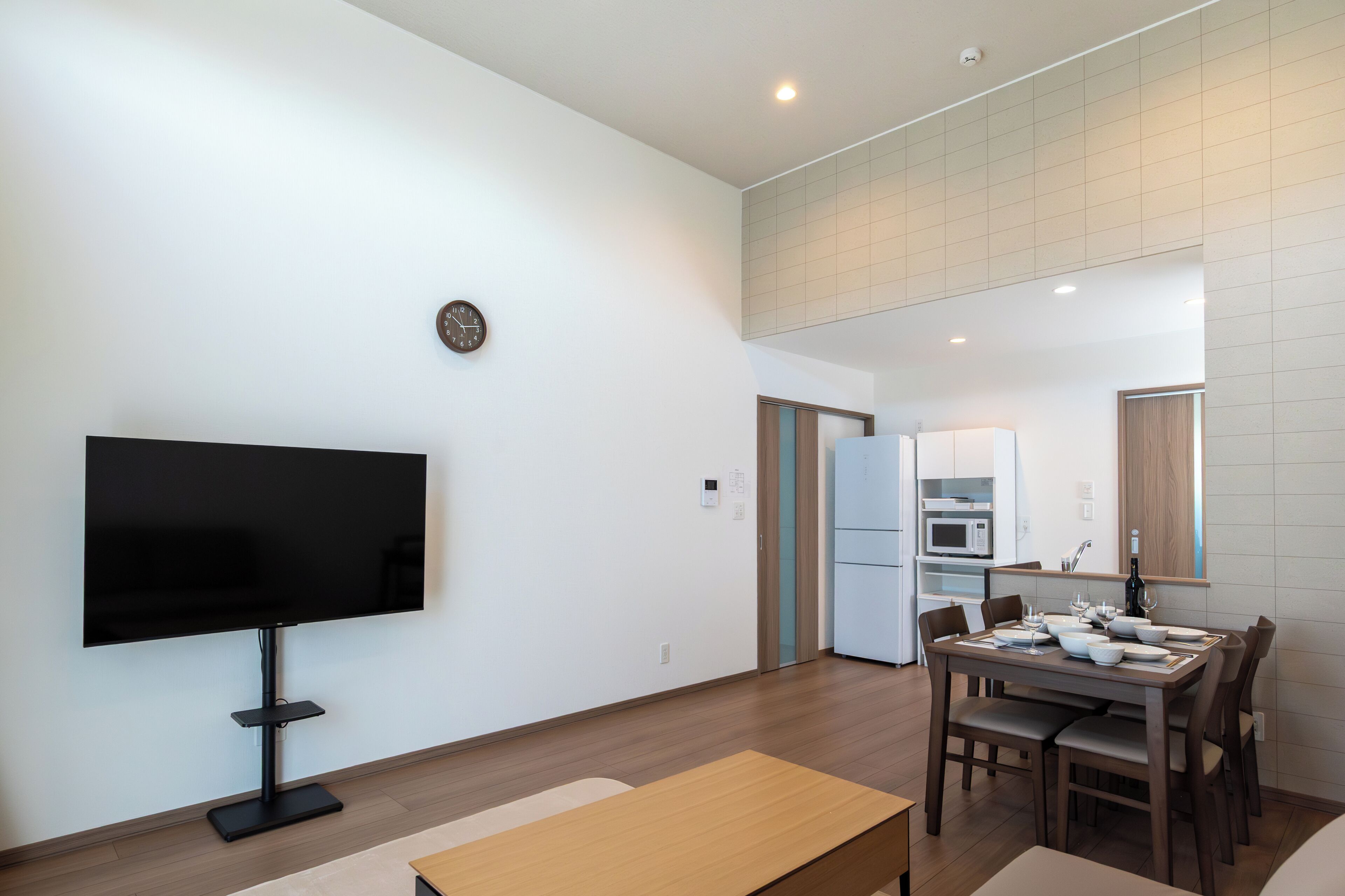 A modern living room with a flat-screen TV, a dining table set for four, and a kitchen in the background.