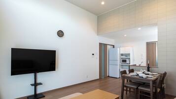 A modern living room with a flat-screen TV, a dining table set for four, and a kitchen in the background.