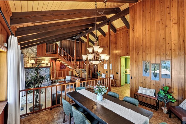 A spacious interior with a high ceiling, wooden beams, a dining area with a long table, and a staircase leading to an upper level.
