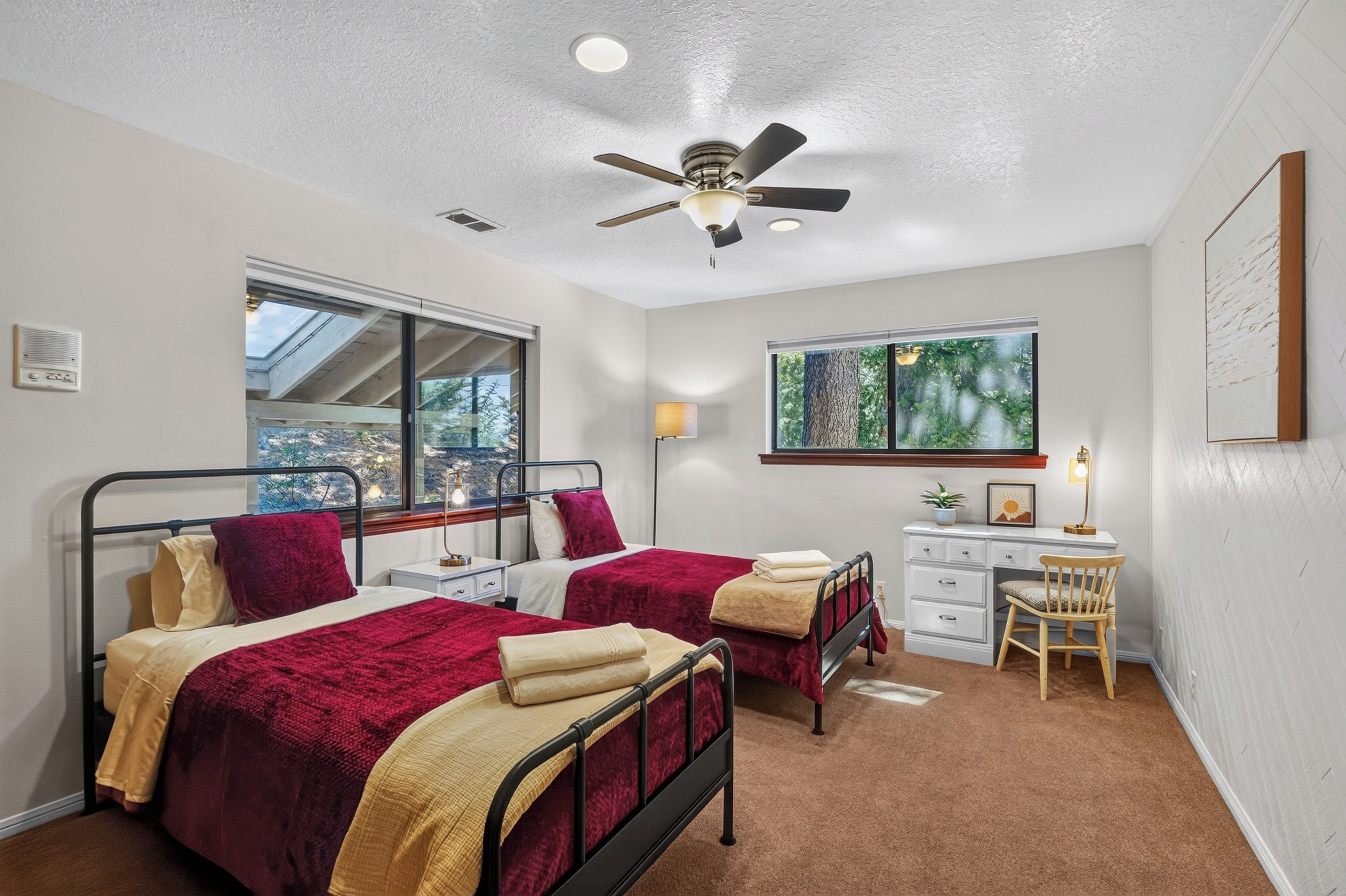 A bedroom with two beds, a desk, a chair, a ceiling fan, and a window with a view of trees.