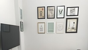 Uma sala de estar moderna com um sofá cinzento, uma TV de tela plana fixada na parede e uma pequena mesa lateral branca com um livro e um abajur.
