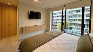 Ein modernes Schlafzimmer mit einem großen Bett, einem angebrachten Fernseher und Blick auf ein Wohngebäude.