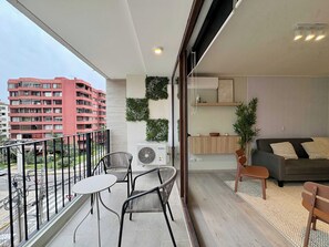 Un balcón con una mesa y sillas, vista de un edificio rojo y una sala de estar con sofá y cojines.