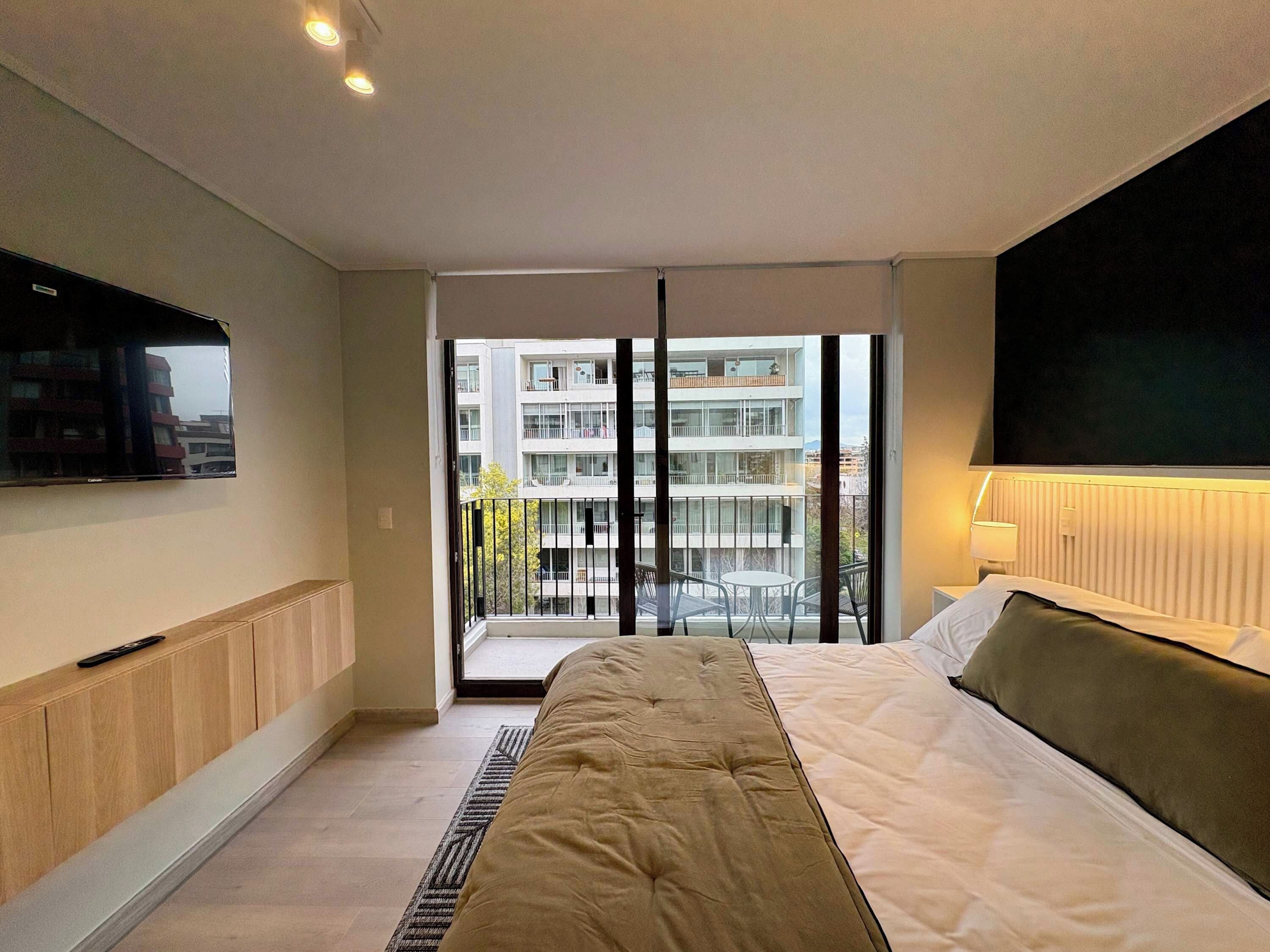 A modern hotel room with a bed, a TV, a balcony with a view of other buildings, and a window.