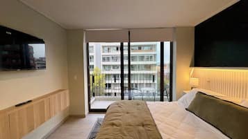 A modern hotel room with a bed, a TV, a balcony with a view of other buildings, and a window.