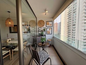 A balcony with a dining area, wicker furniture, and framed wall decorations. - 2BR Las Condes - Parking, Indoor Pool & Laundry (Las Condes)