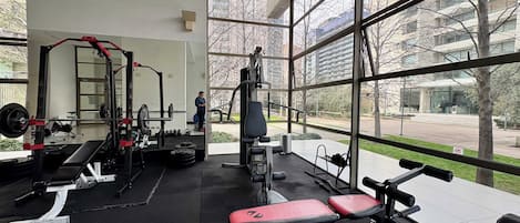 Une salle de sport équipée de divers appareils d’exercice, dotée de grandes fenêtres et offrant une vue sur un immeuble et des arbres.