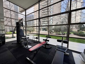 A gym with exercise equipment and large windows overlooking a park and buildings. - 2BR Las Condes - Parking, Indoor Pool & Laundry (Las Condes)