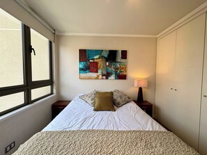 A bedroom with a bed, a window, a nightstand, a lamp, and a wall-mounted painting. - 2BR Las Condes - Parking, Indoor Pool & Laundry (Las Condes)