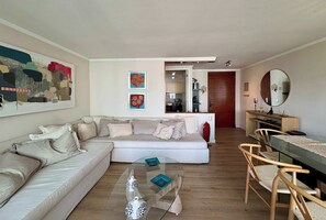 A living room with a sofa, coffee table, and a dining area with chairs and a table. There is a kitchen visible through an open door. - 2BR Las Condes - Parking, Indoor Pool & Laundry (Las Condes)