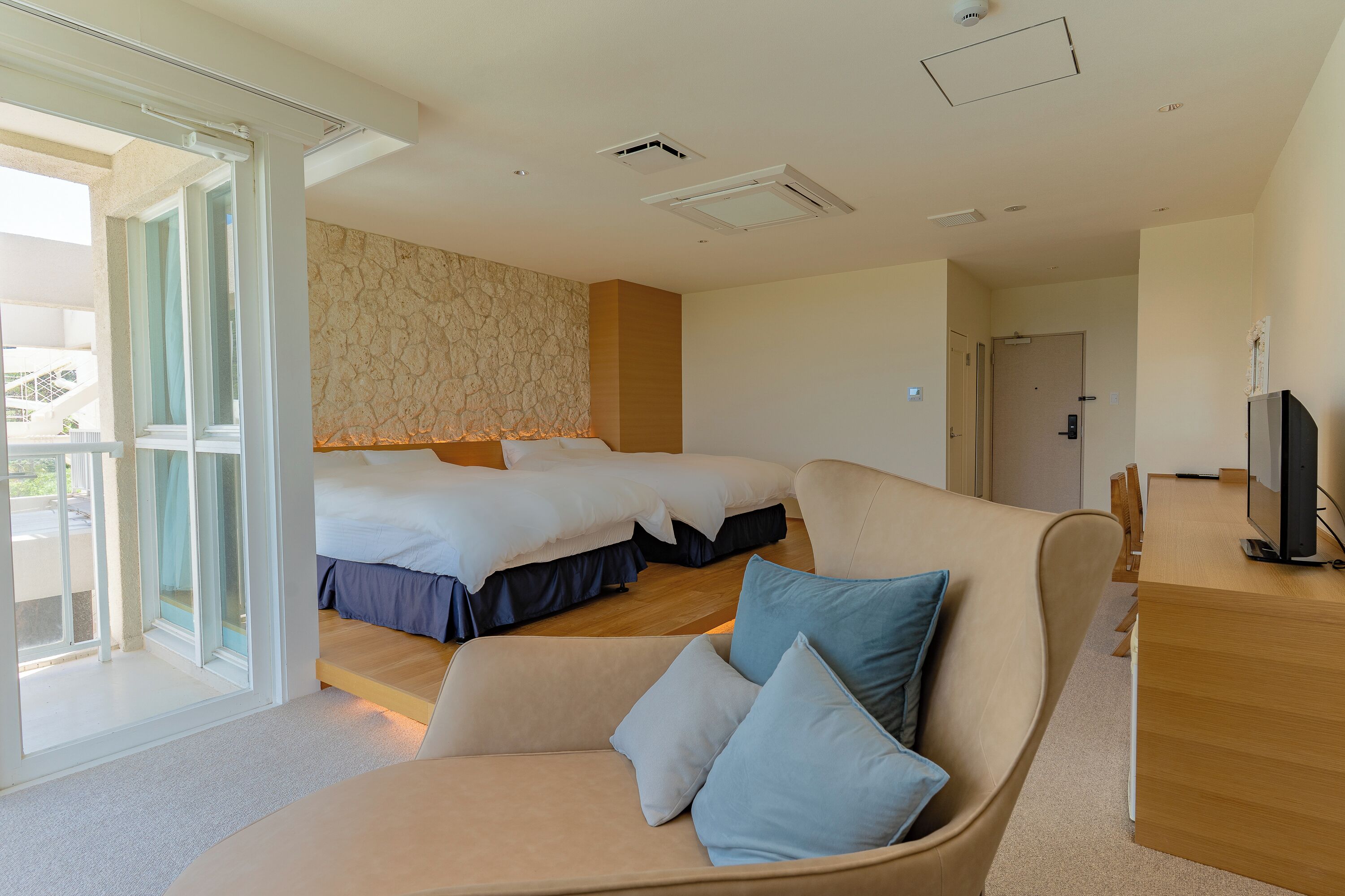 A hotel room with two beds, a sofa, a TV, and a sliding glass door to the outside.