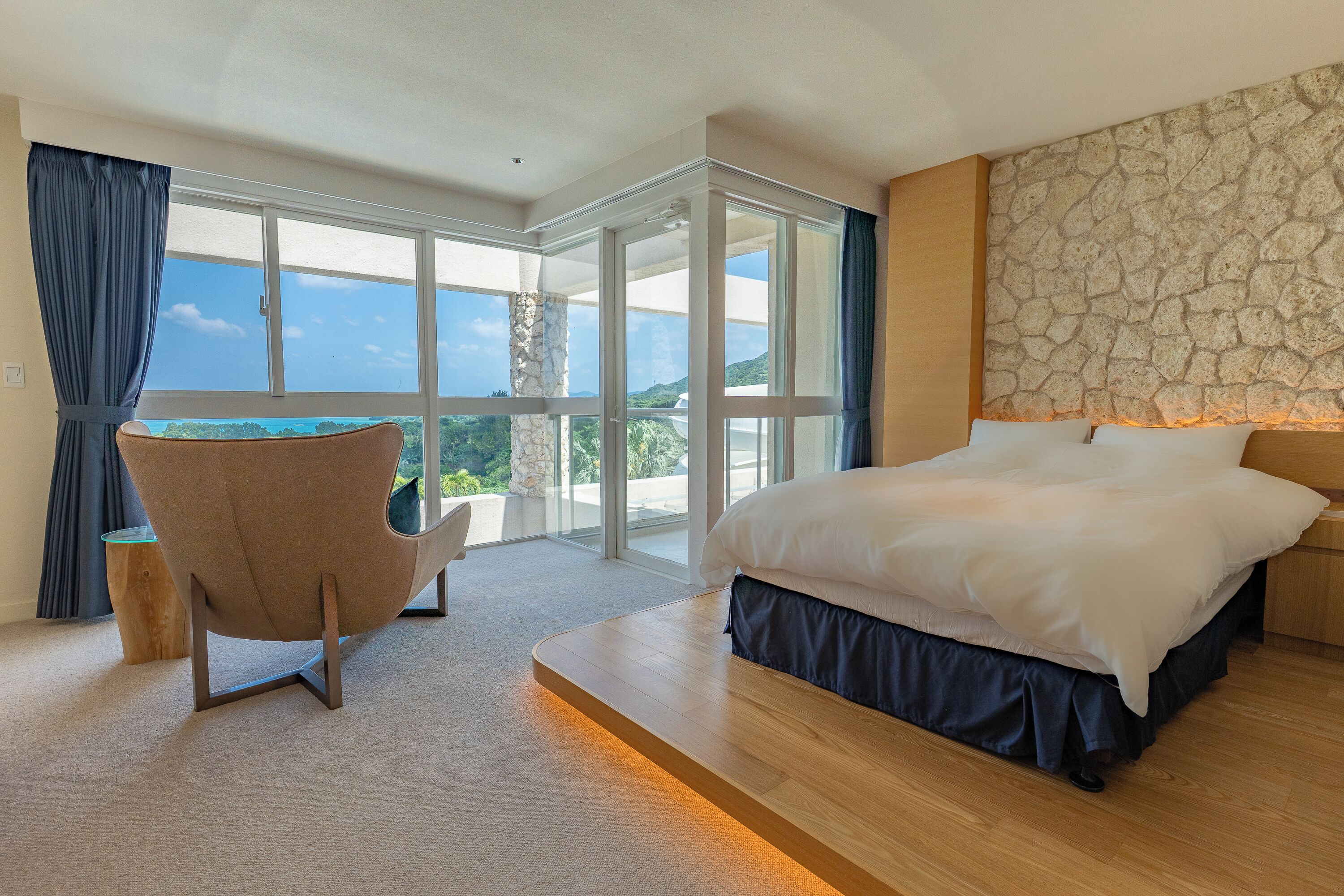 A bedroom with a large bed, a chair, a stone wall, and a view through a glass door.