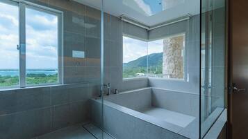 A modern bathroom with a glass-enclosed shower, a bathtub, and large windows offering a view of the sea and mountains.