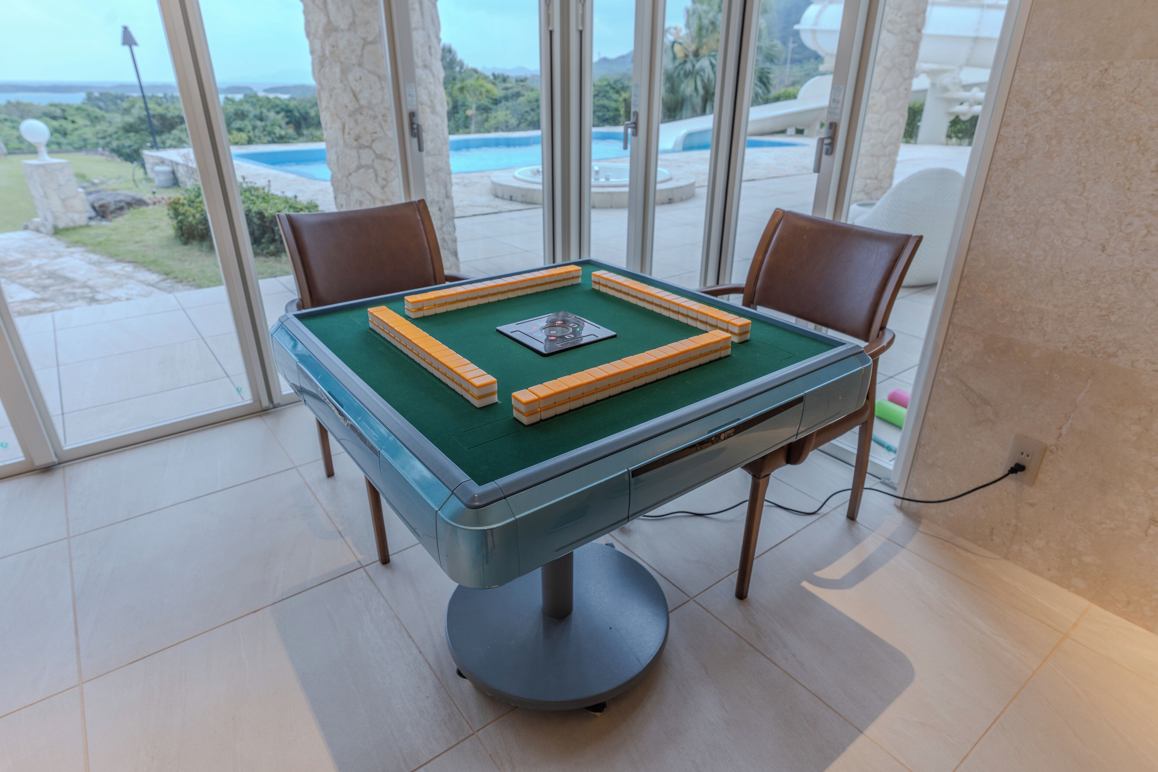 A pool table with a green felt surface, surrounded by chairs, in a room with large windows offering a view of a pool and greenery outside.