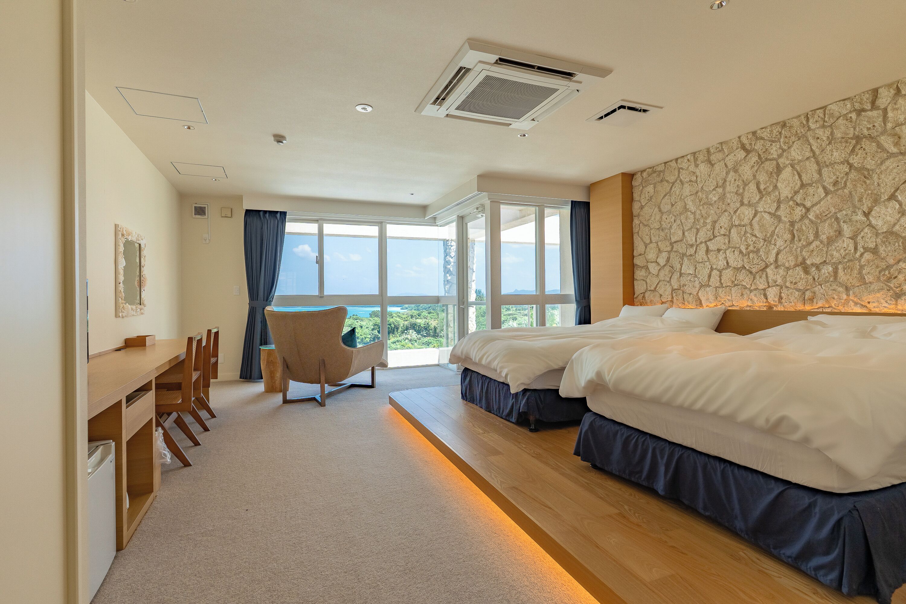 A hotel room with a large bed, a desk, and a chair. There is a view of the outdoors through the windows.